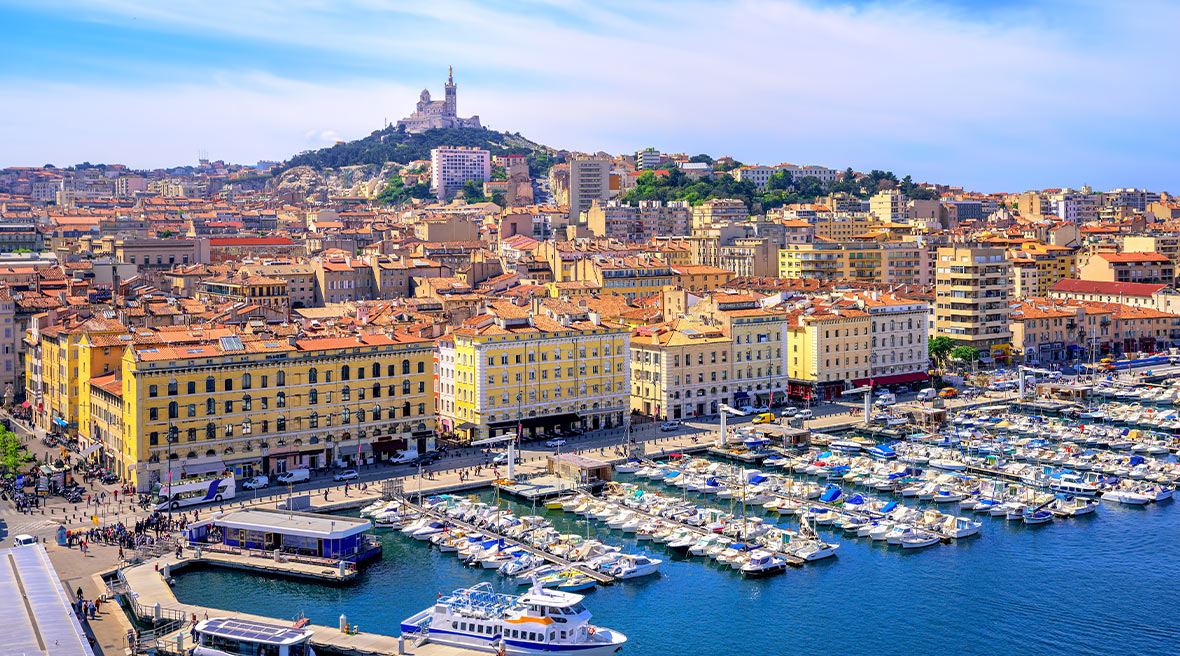 A panoramic view of a large harbour filled with boats and a town of colourful buildings in the distance in the sun